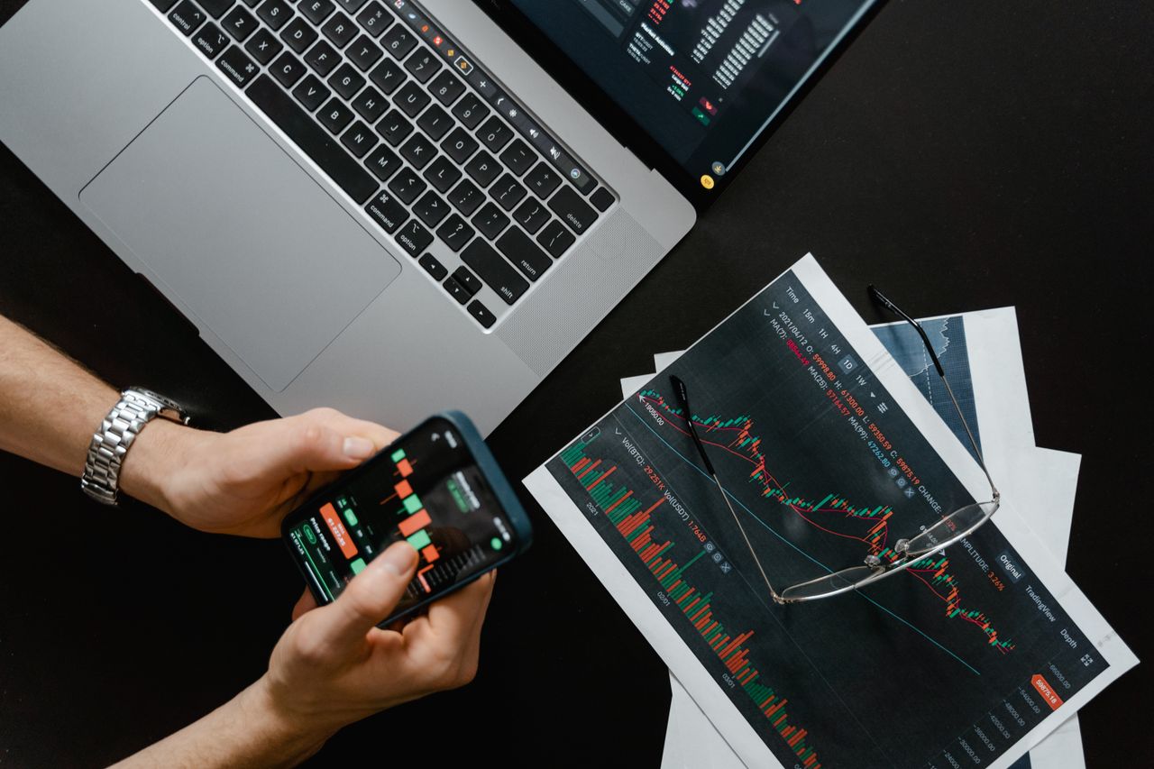 Hands holding a smartphone next to a laptop displaying stock charts, illustrating digital trading.